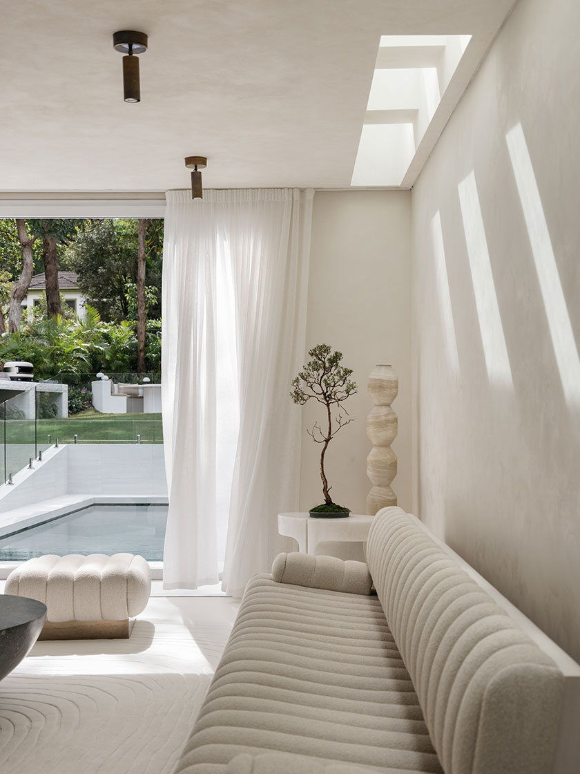 Pavilion – Living A bright living space by Nina Maya Interiors with a view of a sunlit pool, featuring a ribbed white sofa, a sculptural accent table with a bonsai tree, and soft natural shadows cast through a narrow skylight. The overall mood is calm and contemplative, accentuated by clean architectural lines.