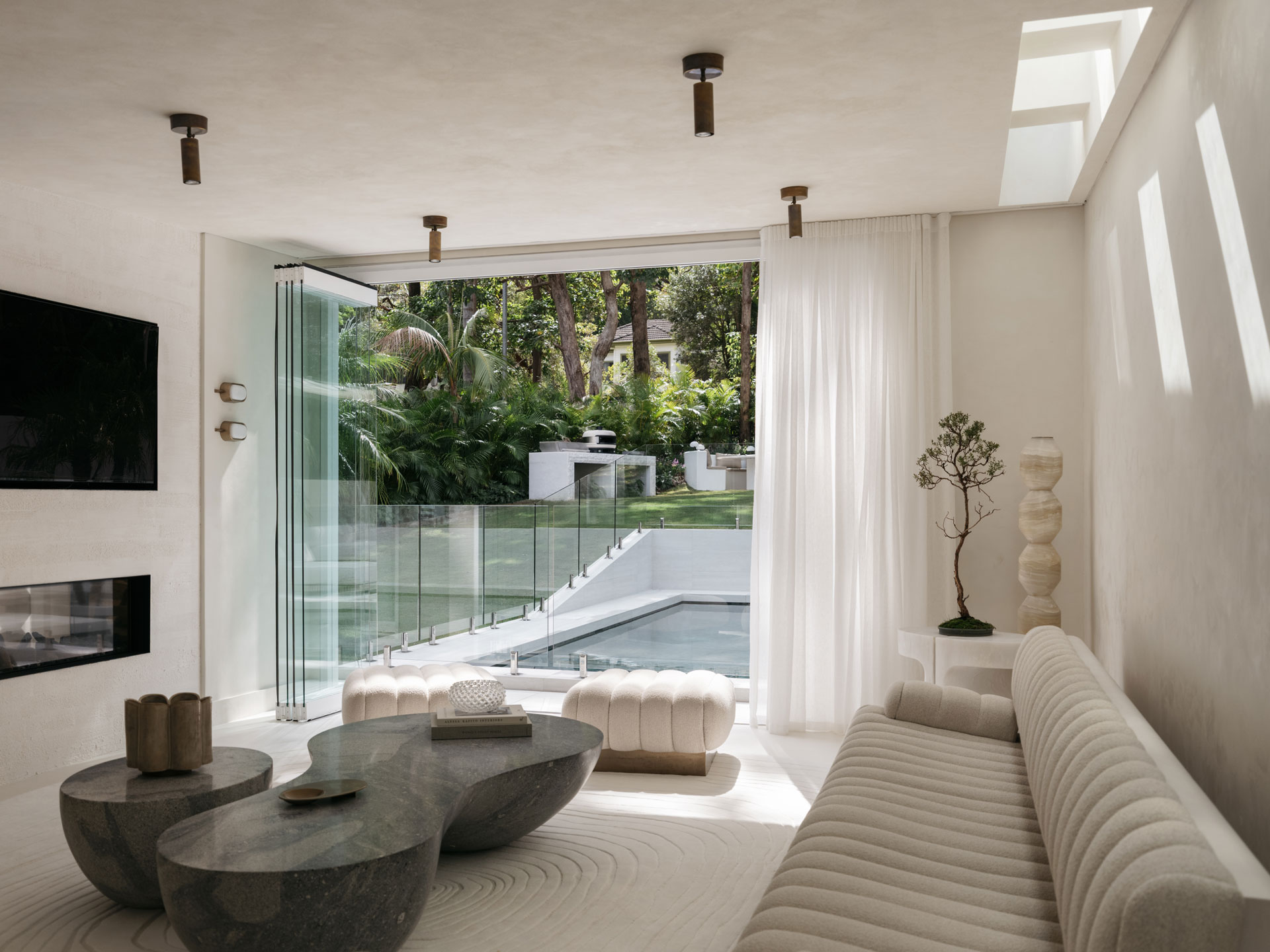 Pavilion – indoo-outdoor-connection A contemporary living space by Nina Maya Interiors, where sculptural stone coffee tables and a ribbed boucle sofa sit beside full-height glass doors opening to a lush garden and pool. The soft natural light, sheer curtains, and bonsai accent create a calm indoor-outdoor connection.