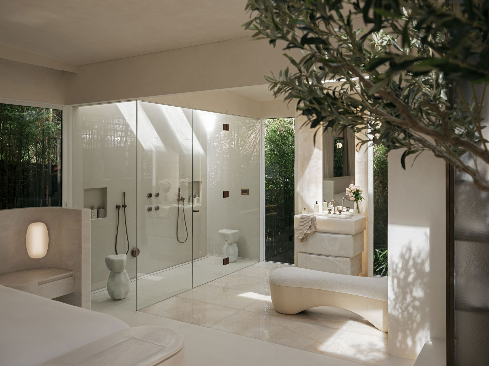 Avalon – bathroom An ethereal bathroom scene by Nina Maya Interiors with a large glass shower enclosure, a carved stone vanity, and a sculptural bench nestled beneath an olive tree. Surrounded by lush greenery and bathed in natural light, the room embodies a tranquil, spa-like ambiance.