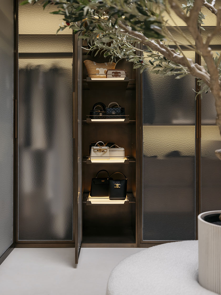 Pavilion – WIR A curated wardrobe nook designed by Nina Maya Interiors, featuring a bronze-framed glass cabinet displaying a collection of luxury handbags, including Chanel, Hermès, and Celine, all lit by soft interior lighting. An olive tree partially frames the open shelving, enhancing the organic elegance.