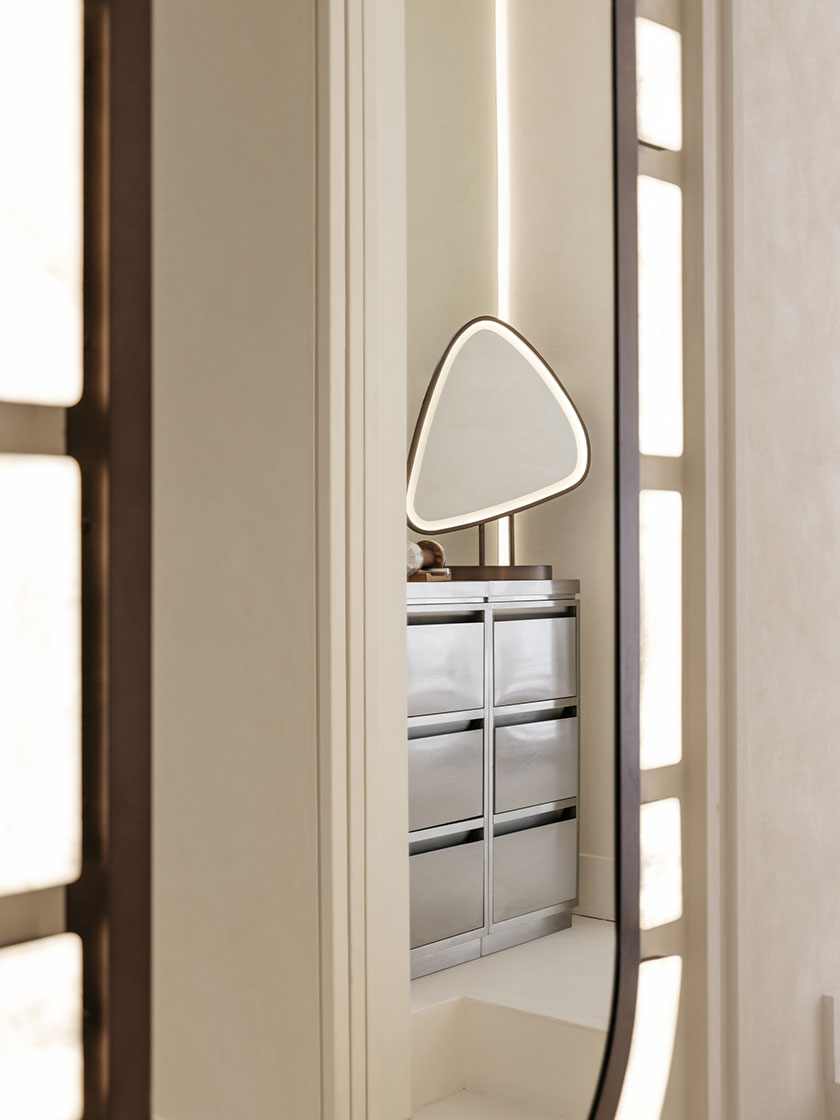 Pavilon – Dressing area A softly lit dressing area by Nina Maya Interiors, viewed through a glowing oval mirror. The space includes a metallic drawer console and a modern triangular vanity mirror, bathed in warm reflected light that highlights the room’s textural minimalism.