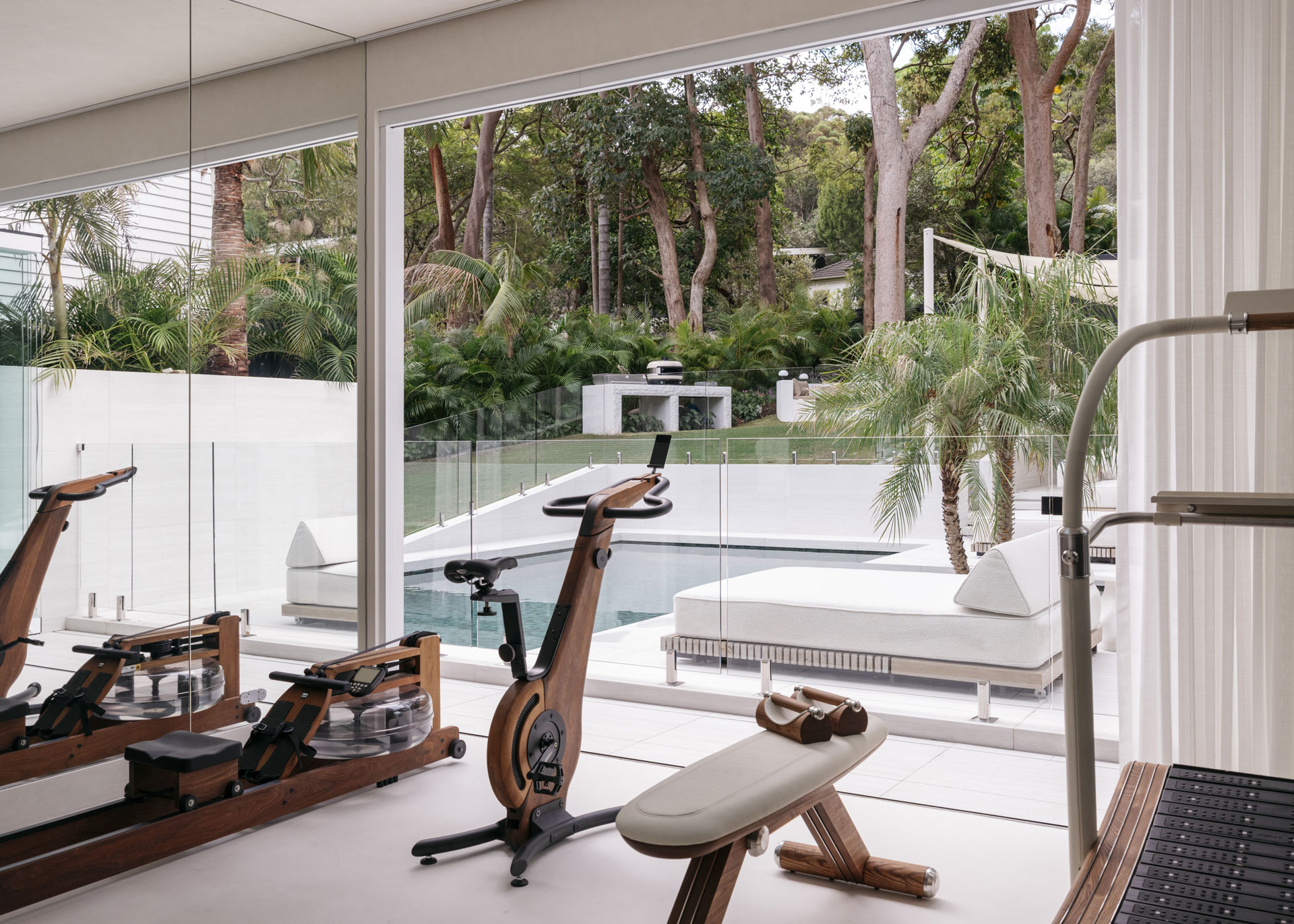 A sleek home gym by Nina Maya Interiors opens directly onto a tranquil poolside area, with full-height glass doors providing uninterrupted views of palm trees and manicured greenery. Natural light fills the space, which features a wooden water rower, a bike, and a sculptural bench.