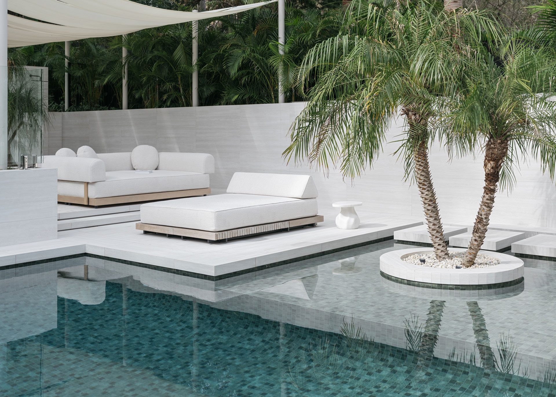 A peaceful outdoor lounge designed by Nina Maya Interiors, showcasing white minimalist daybeds and circular armrest sofas beside a tiled pool. Surrounded by tropical palms and a stone privacy wall, the space is shaded by a soft canopy and grounded by sculptural white accent tables.