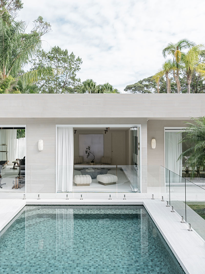 Pavilion – rear exterior A rear exterior view of the home designed by Nina Maya Interiors, with full-width sliding doors opening from the living room onto a green-tiled pool. Inside, curved ivory furniture and a bonsai artwork are visible, blending interior elegance with the lush garden surroundings.