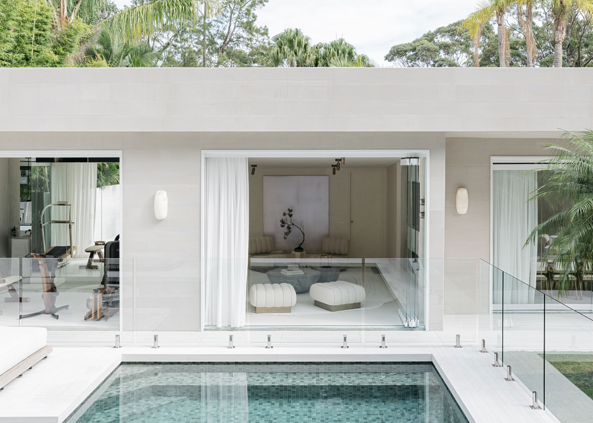 A wider perspective of the home's rear façade by Nina Maya Interiors, where two minimalist interior spaces open onto a sleek pool with glass fencing. The neutral stone cladding and modern silhouette enhance the seamless flow between indoor serenity and tropical outdoor beauty.