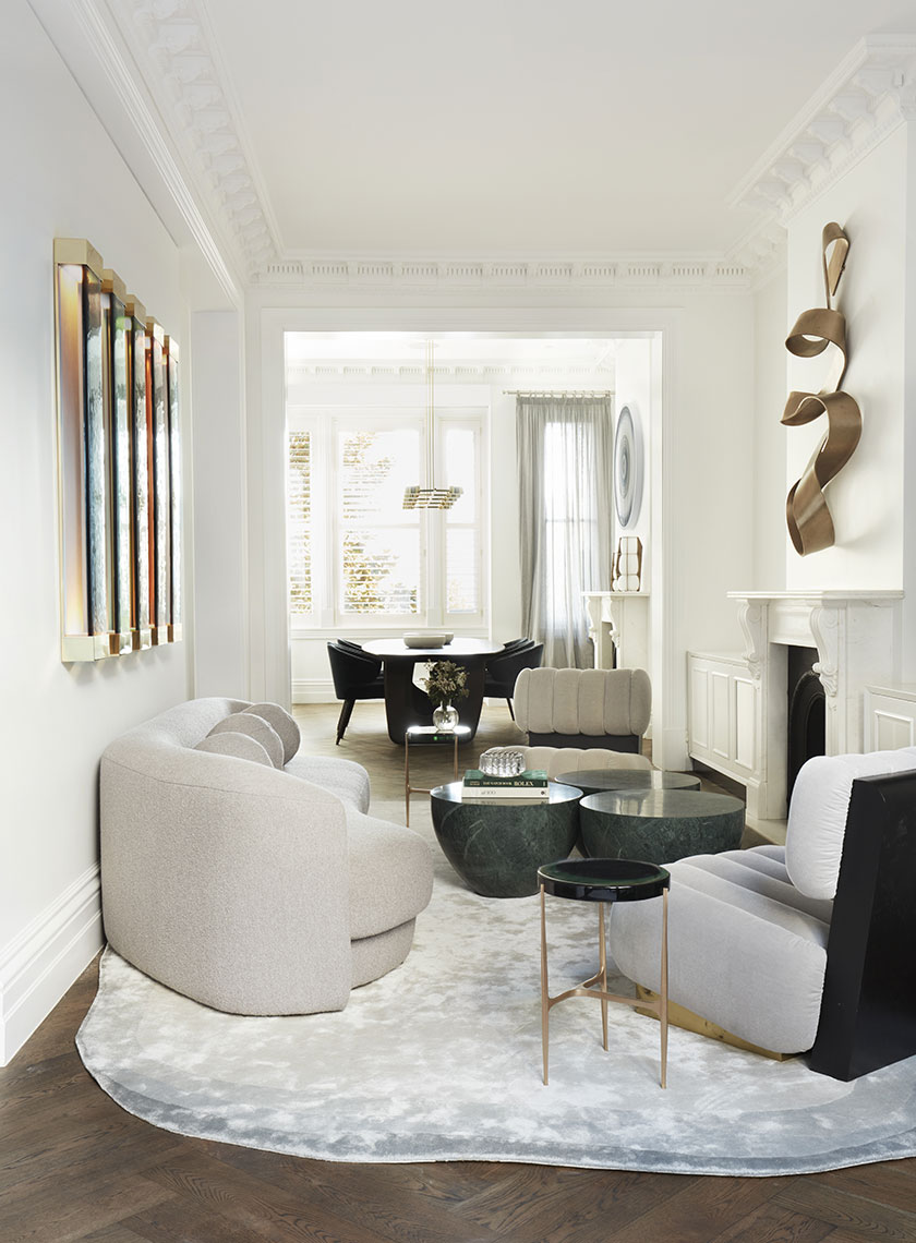 19_Nina-Maya-Residence_K A sophisticated living room with curved sofas, green marble coffee tables, and sculptural bronze wall art, leading into a bright dining area with large windows. Interior design by Nina Maya Interiors.