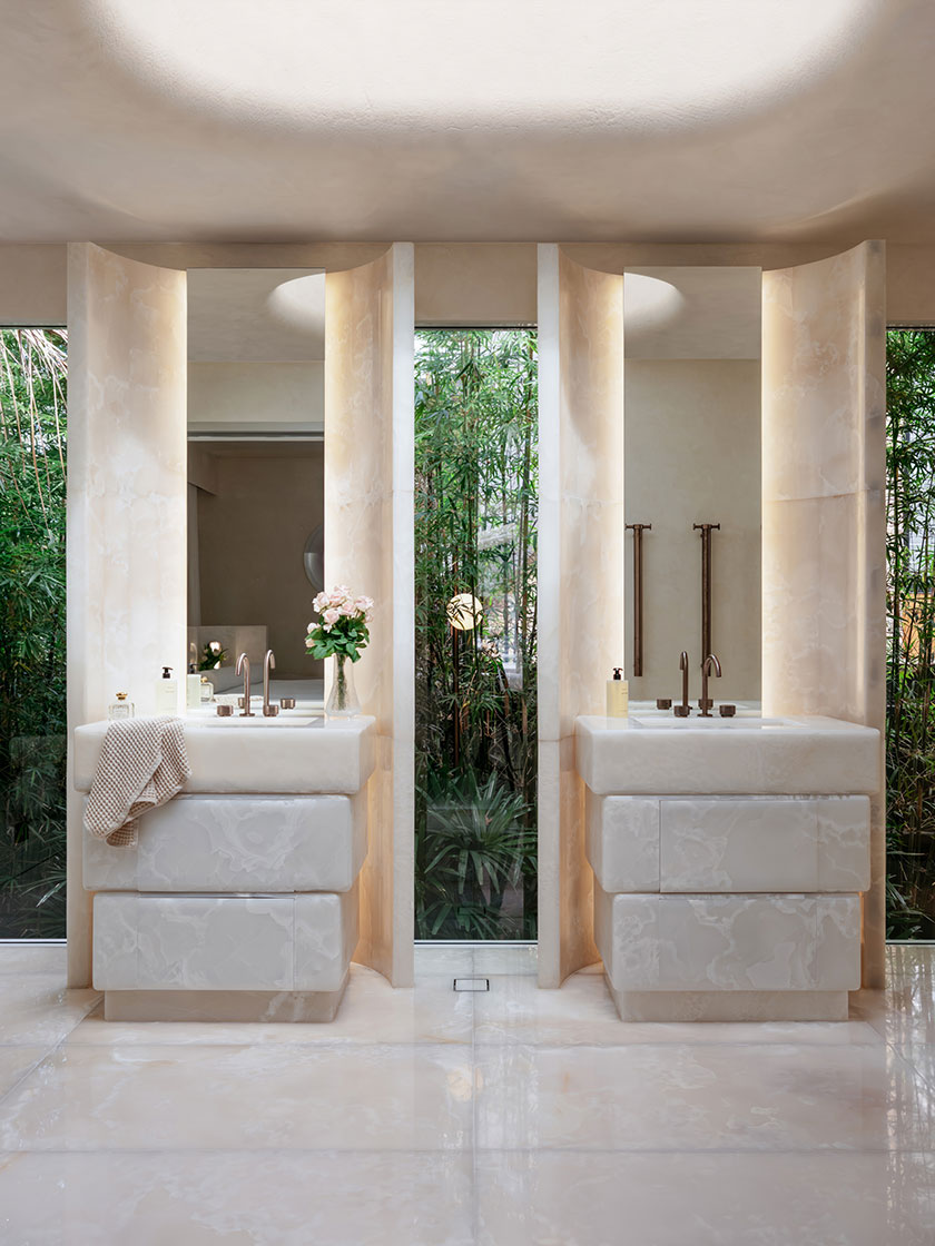 2 A luxurious double vanity bathroom by Nina Maya Interiors, showcasing two carved stone sinks framed by vertical onyx panels and floor-to-ceiling windows overlooking bamboo greenery. Bronze fixtures, soft uplighting, and floral arrangements add refined touches to the spa-like atmosphere.