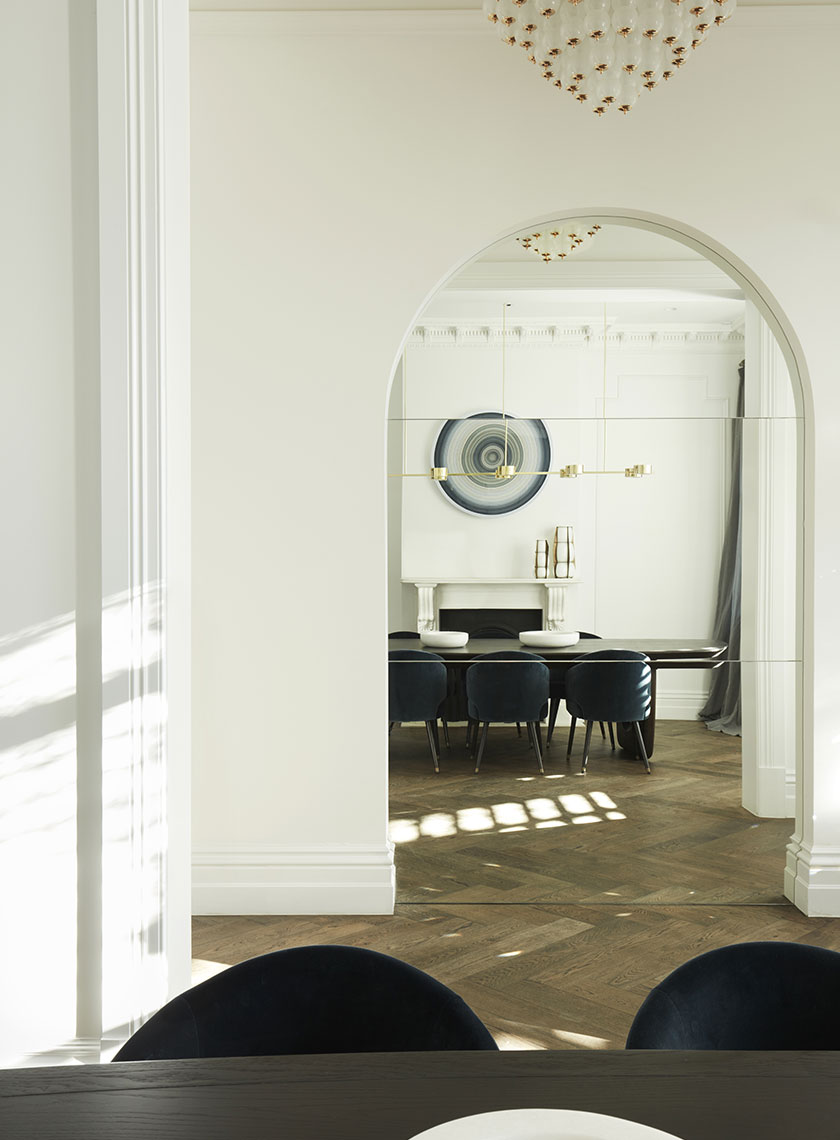 21_Nina-Maya-Residence_K A formal dining room viewed through a large archway, featuring a modern brass chandelier, dark wood dining table, and a circular piece of art on the wall. Interior design by Nina Maya Interiors.