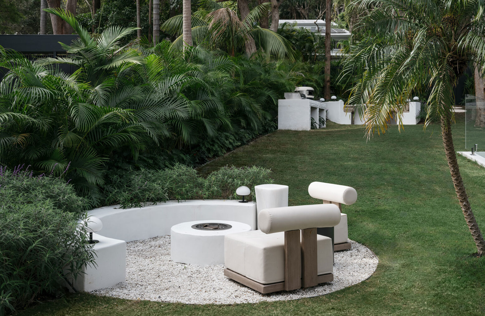 Pavilion fire pit A sculpted fire pit lounge by Nina Maya Interiors nestled in a circular gravel zone, surrounded by palm trees and dense garden foliage. The space features curved white bench seating, cylindrical cushions, and modern outdoor lighting for a cozy, organic gathering n