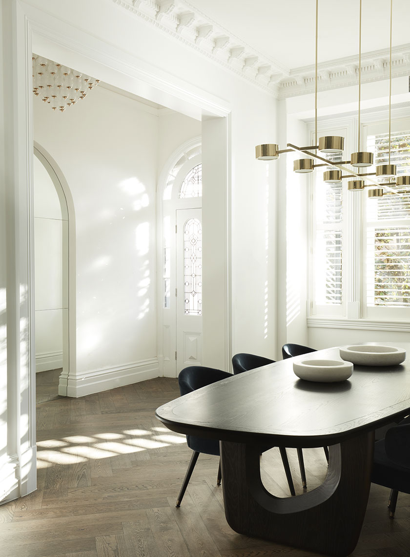 22_Nina-Maya-Residence_K A dining room with a large, dark wood table surrounded by blue velvet chairs, illuminated by a contemporary brass chandelier under a white, detailed ceiling. Interior design by Nina Maya Interiors.