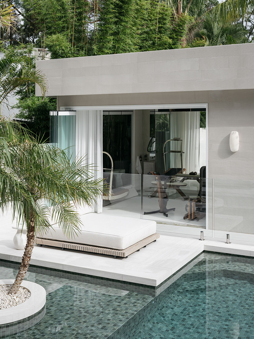 Pavilion – poolside retreat A tranquil view of a Nina Maya Interiors poolside retreat, with a low white daybed set beneath tropical palms and lush bamboo. The adjacent gym is visible through glass doors, seamlessly blending wellness and relaxation.