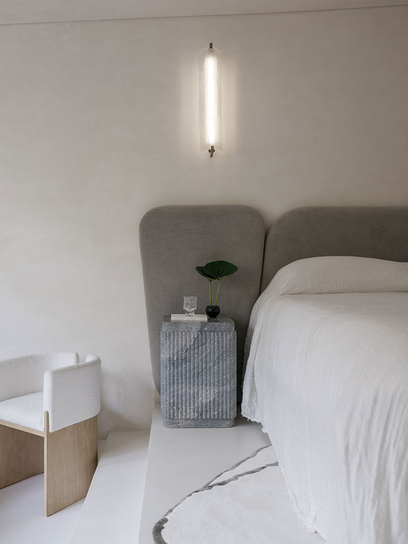 Pavilion – bedroom A calming bedroom corner by Nina Maya Interiors, featuring a sculptural grey headboard, a ribbed stone nightstand, and a softly glowing wall sconce. Natural textures are balanced by a boucle chair and minimalist styling.