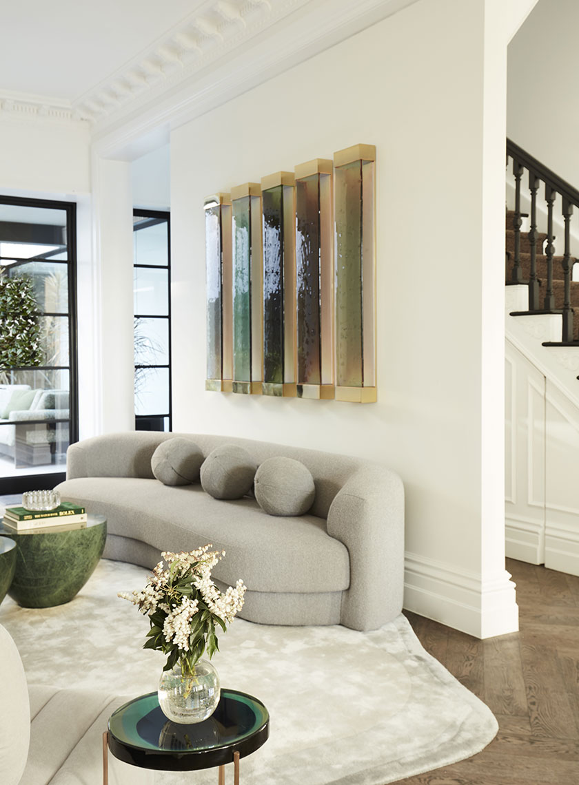25_Nina-Maya-Residence_K A close-up of a curved gray sofa with round cushions, positioned in front of modern, backlit wall art in warm tones. Interior design by Nina Maya Interiors.
