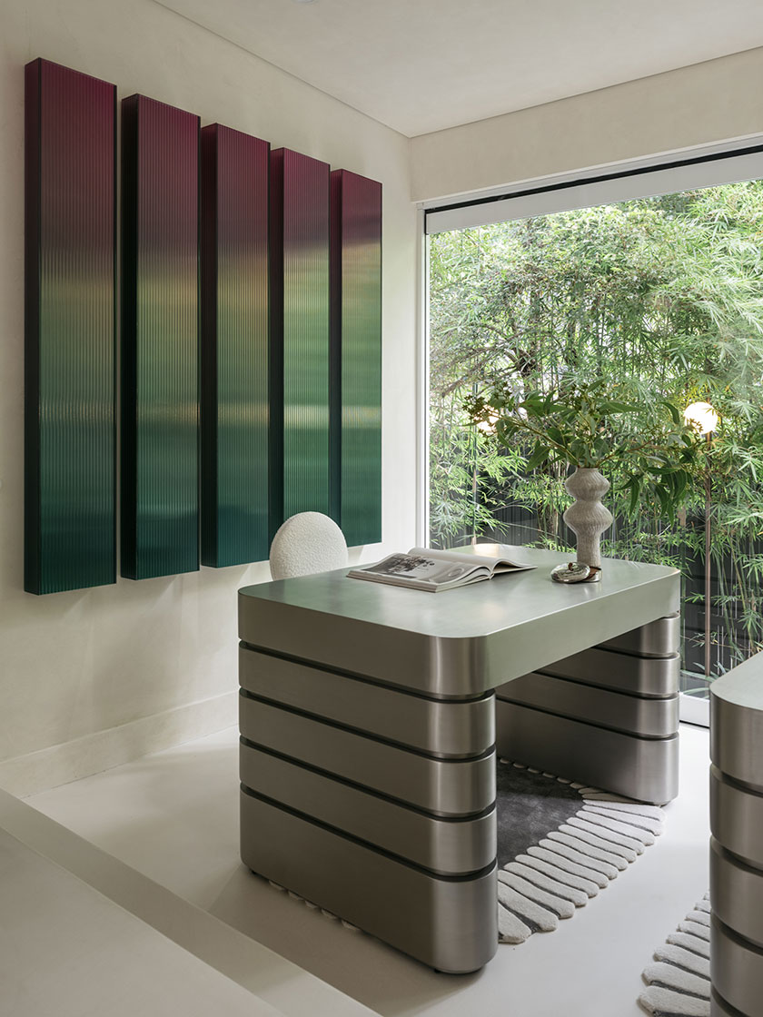 Pavilion – Study nook A bold study nook designed by Nina Maya Interiors, featuring a brushed stainless steel desk with horizontal grooves and an ombré gradient wall installation in burgundy and green tones. Floor-to-ceiling windows open to dense bamboo, with a sculptural vase and neutral rug grounding the space.