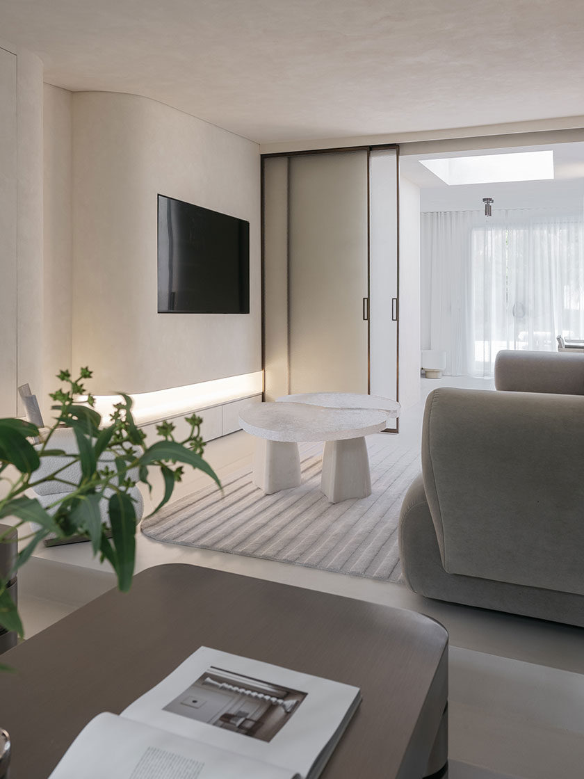 Pavilion – TV lounge A softly lit TV lounge by Nina Maya Interiors with curved taupe sofas, a sculptural white coffee table on a ribbed cream rug, and frosted sliding glass doors leading to a bright atrium. The minimal design is softened with greenery and tactile surfaces.