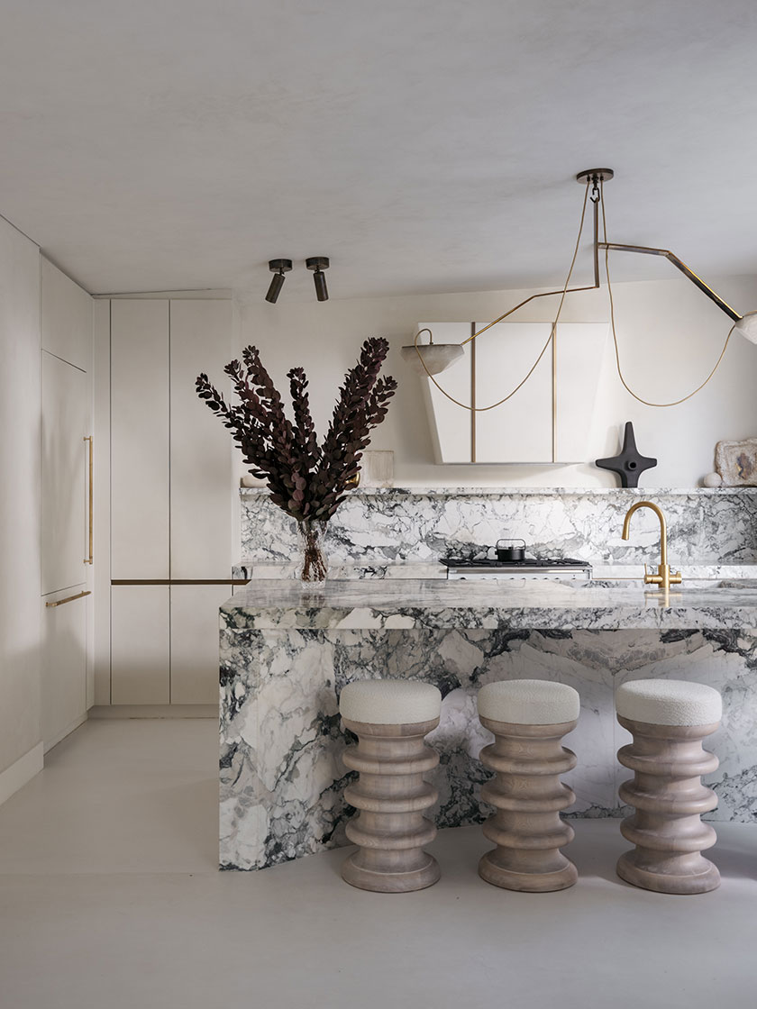 Pavilion – Kitchen A striking kitchen designed by Nina Maya Interiors with dramatic marble surfacing across the island and splashback, paired with sculptural timber stools topped in boucle. Brass pendant lighting and gold-toned fixtures add warmth and contrast to the cool-toned palette.