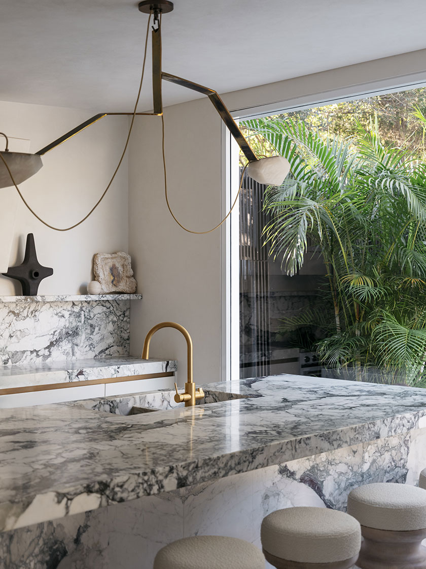 Pavilion – Kitchen island A close-up kitchen view by Nina Maya Interiors with a sculptural brass faucet, rich marble counters, and tropical greenery just outside the window. The layered pendant light fixture casts soft shadows on the ceiling, enhancing the organic elegance of the space.