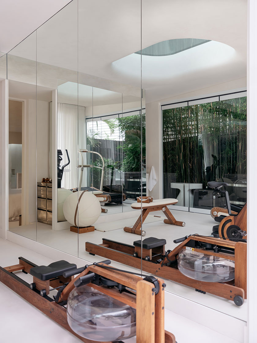Pavilion – Gym A serene home gym by Nina Maya Interiors, reflecting soft natural light through mirrored walls and a curved skylight. Equipment includes a wooden water rowing machine, balance ball, elliptical trainer, and a minimalist bench, all framed by sheer drapery and a bamboo-filled courtyard beyond.