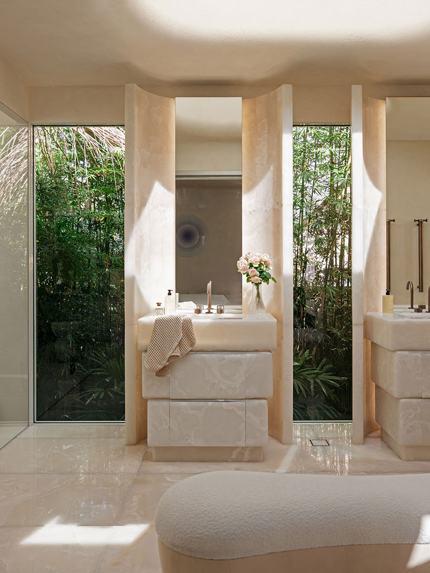 Pavilion -bath A luxurious bathroom by Nina Maya Interiors, featuring a sculpted onyx vanity with fluted vertical accents and rose-filled glass vases. Surrounded by full-height windows revealing lush bamboo, the space is softly lit by a ceiling skylight, with a boucle bench subtly placed in the foreground.