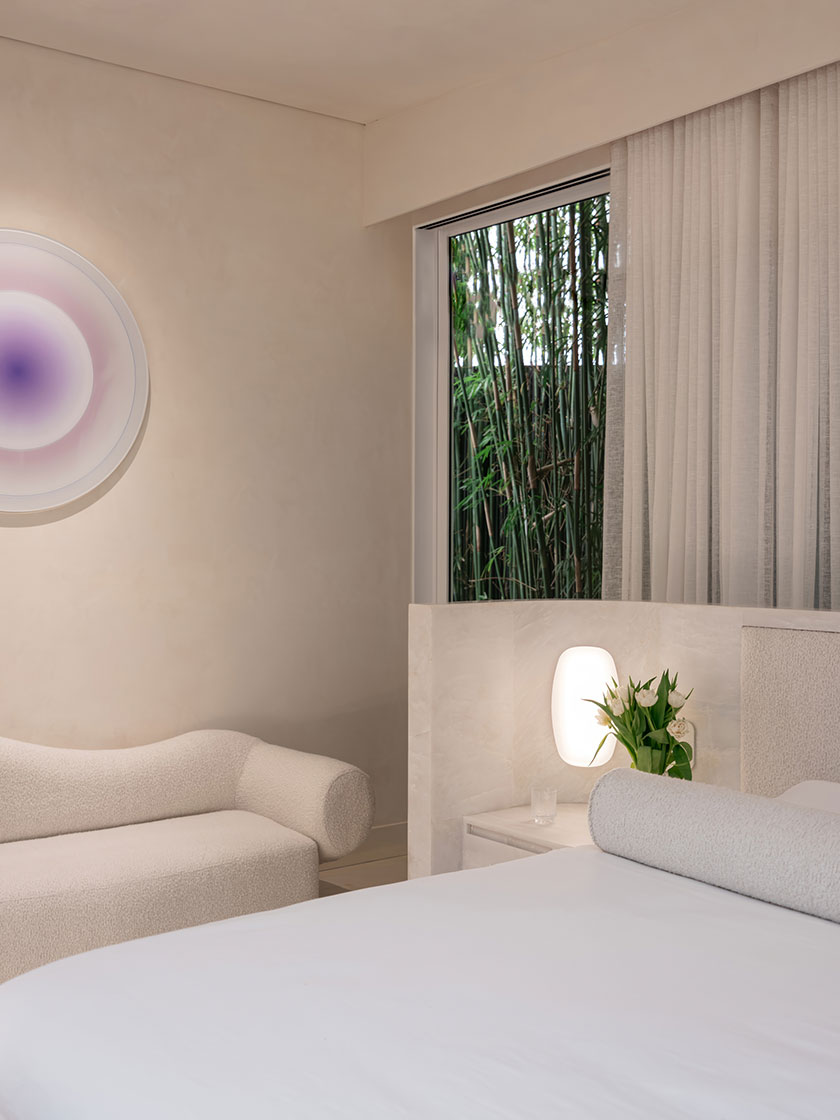Pavilion – bed A serene bedroom corner by Nina Maya Interiors showcasing a plush boucle chaise, crisp white bedding, and a softly glowing sculptural bedside lamp. A round wall-mounted artwork in lavender tones adds a touch of whimsy, while sheer drapes frame a view of dense bamboo outside.