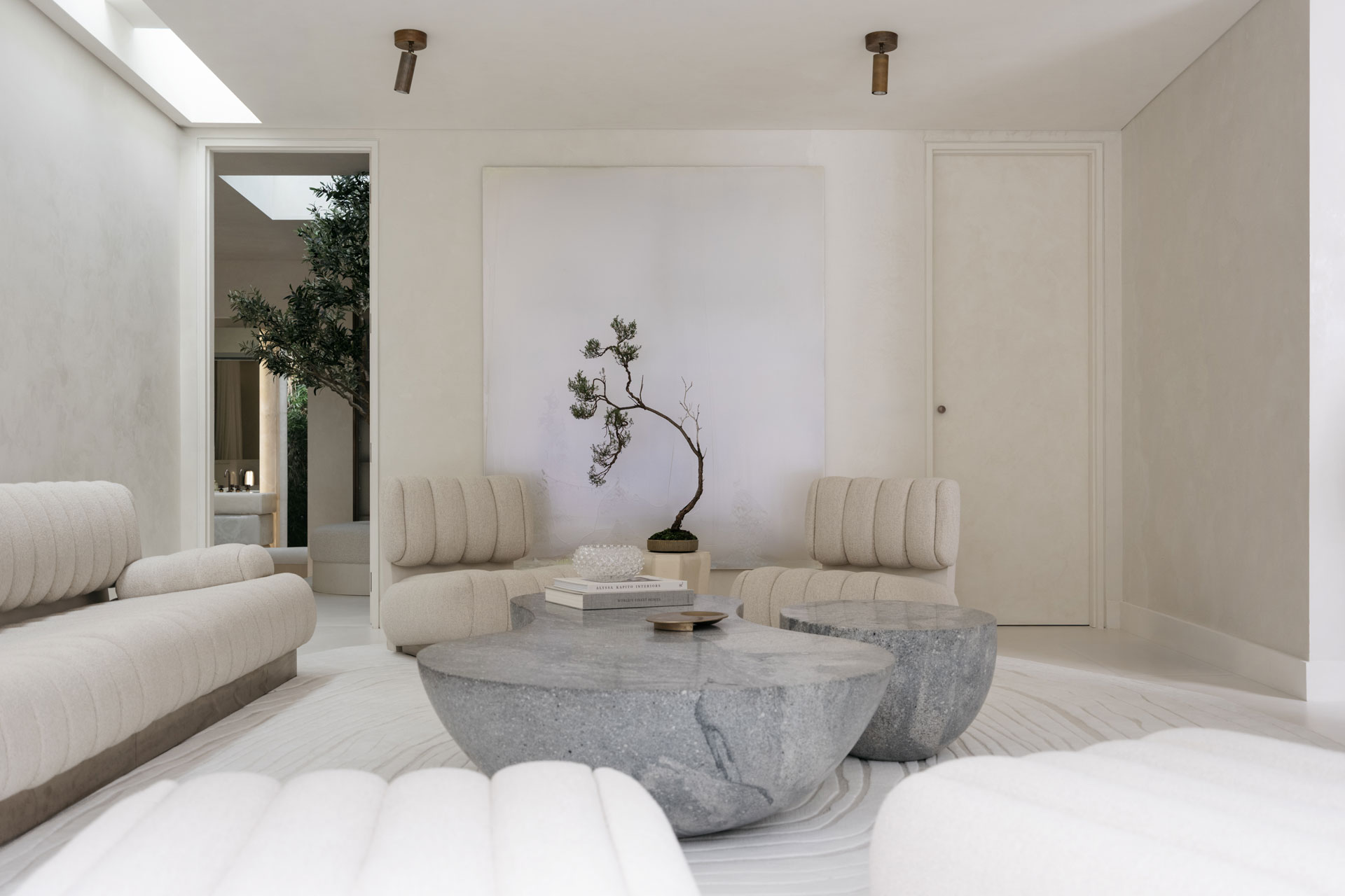 Pavilion – living A minimalist living room by Nina Maya Interiors with channel-tufted ivory seating, sculptural grey stone coffee tables, and a striking bonsai-inspired art piece. The neutral palette is warmed by natural textures and indirect light filtering from a skylight beyond.