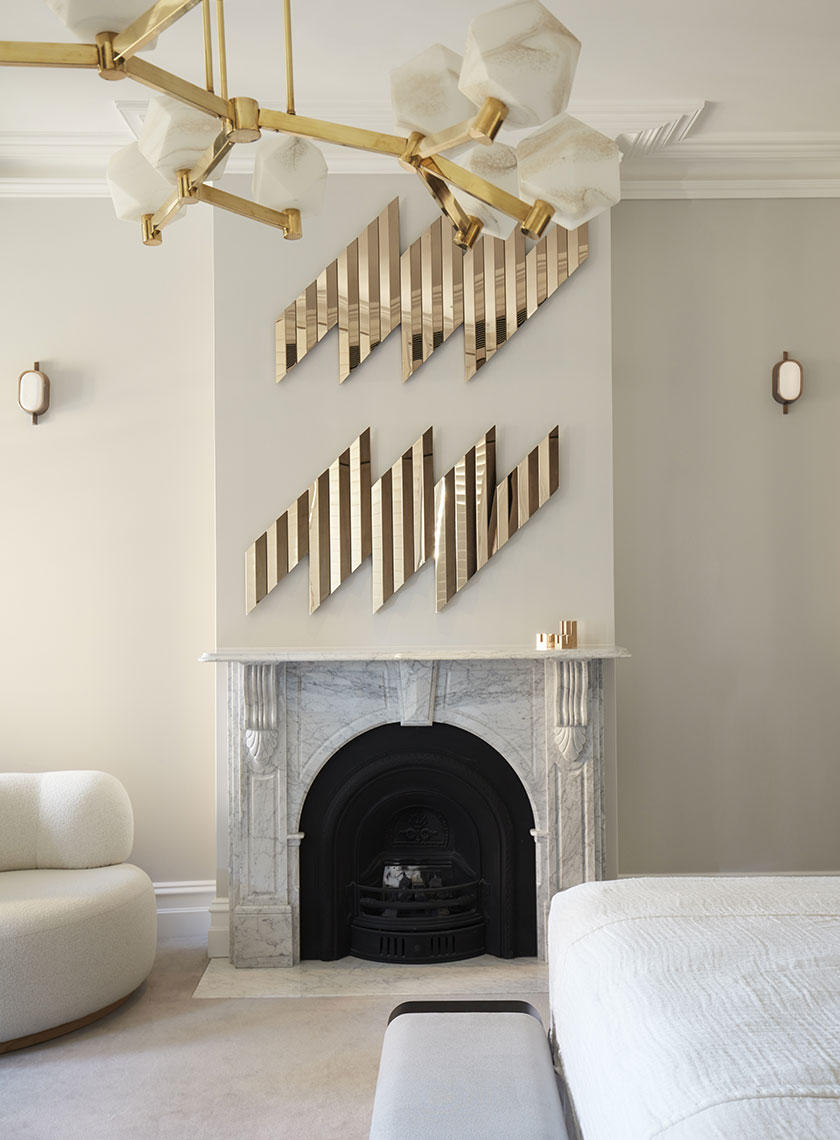 Nina-Maya-Victoria-st2575 A chic fireplace in a modern room, with a marble surround and a black fireplace insert, accented by gold, geometric wall art above. A brass chandelier with hexagonal lights hangs from the ceiling, complementing the overall luxe aesthetic crafted by Nina Maya Interiors. The room is designed with soft, neutral tones, featuring a curved white sofa in the corner.