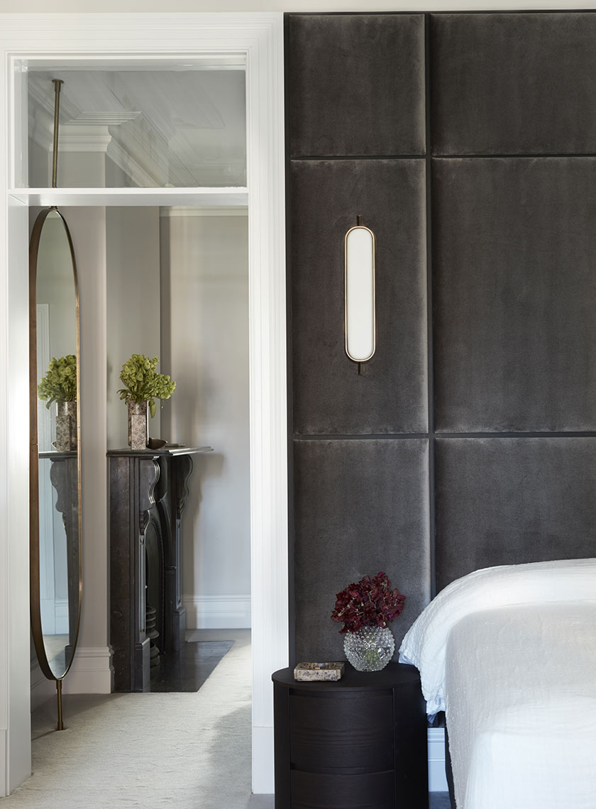 Nina-Maya-Victoria-st2593 A luxurious walk-in wardrobe with dark, glass-fronted wardrobes illuminated by soft interior lighting. A circular grey ottoman with a black handbag sits in the center, beneath a glass-topped side table. The wardrobe opens into a bright room with a marble fireplace, highlighting the seamless blend of functionality and style by Nina Maya Interiors.