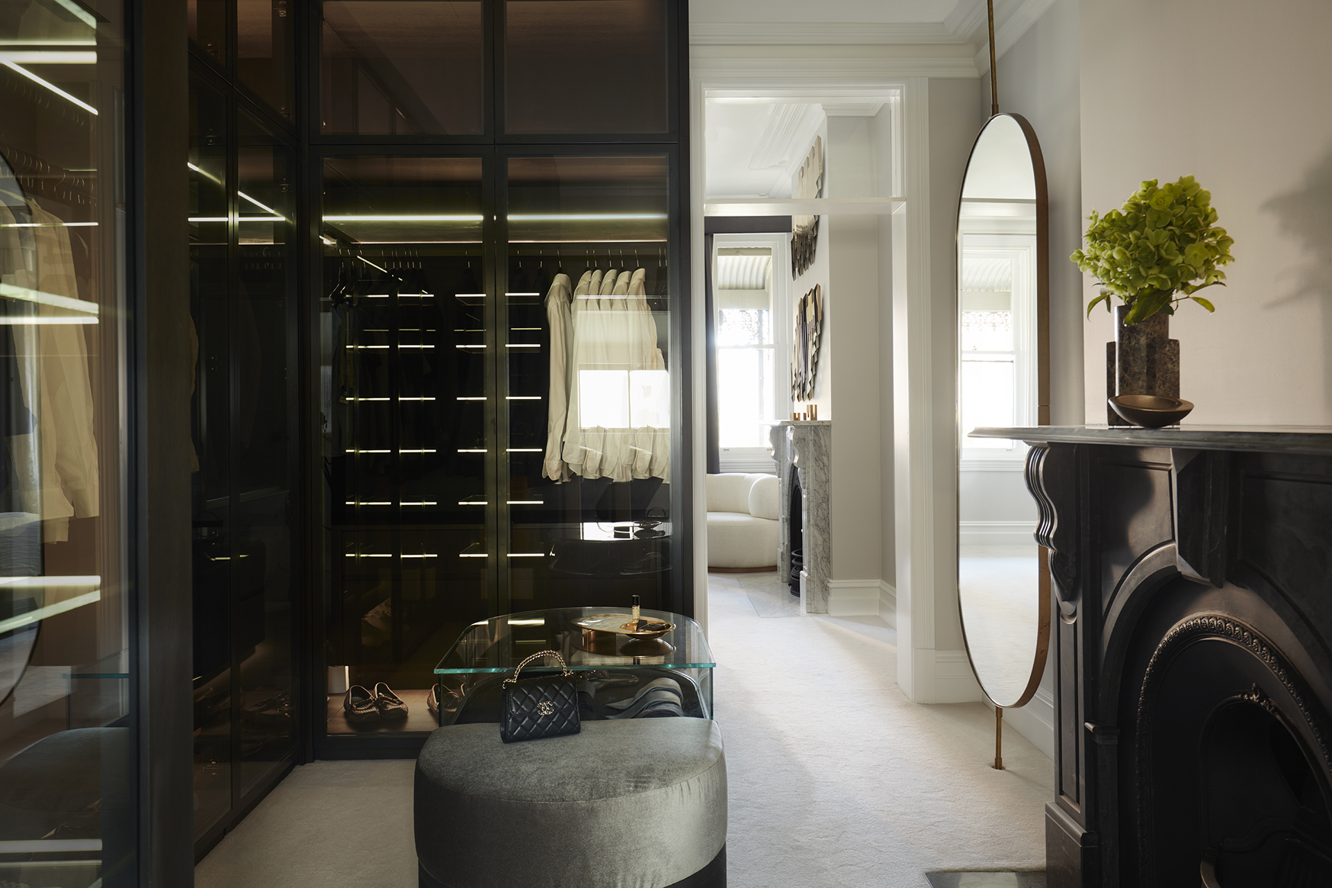 Nina Maya Victoria st2624 Walk-in Closet with Glass Doors: A luxurious walk-in closet featuring glass-paneled wardrobe doors, a plush round ottoman, and a sleek black fireplace, designed by Nina Maya Interiors at Residence K, Potts Point.