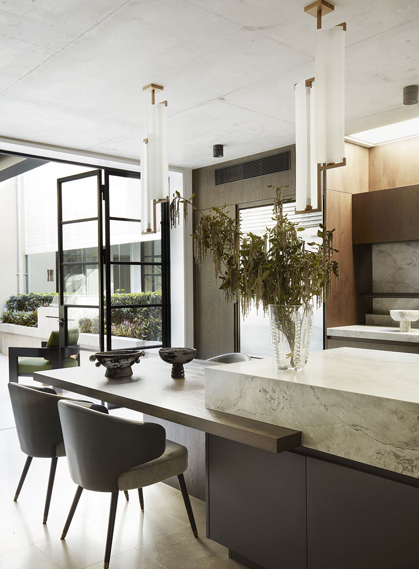Nina-Maya-Victoria-st2762 A modern kitchen and dining area featuring a marble island, wooden cabinetry, and a vase of dried branches on the table, with floor-to-ceiling glass doors opening to an outdoor space. Interior design by Nina Maya Interiors.