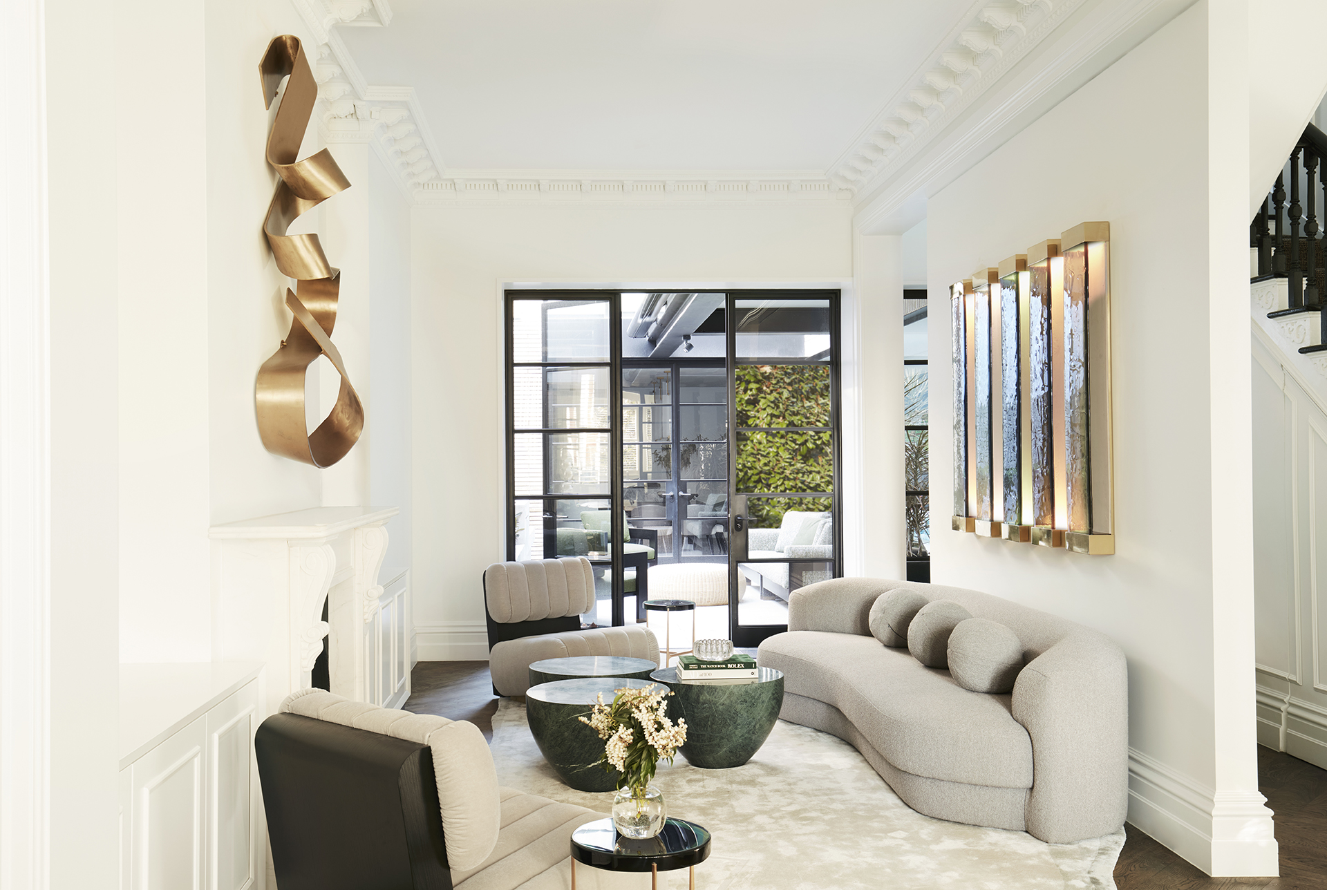 Nina Maya Victoria st2874 2 A stylish living room with a curved gray sofa, green marble coffee tables, and modern art on the walls, with a view of an outdoor seating area through glass doors. Interior design by Nina Maya Interiors.